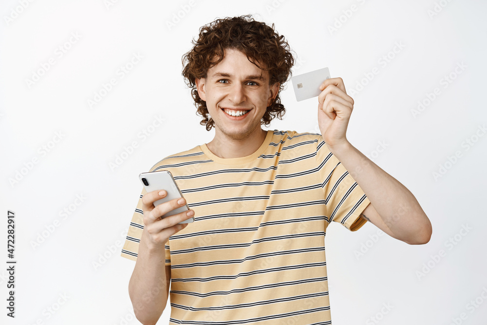 Handsome smiling curly guy showing credit card and smartphone, concept of online shopping, white background Handsome smiling curly guy showing credit card and smartphone, concept of online shopping, white background