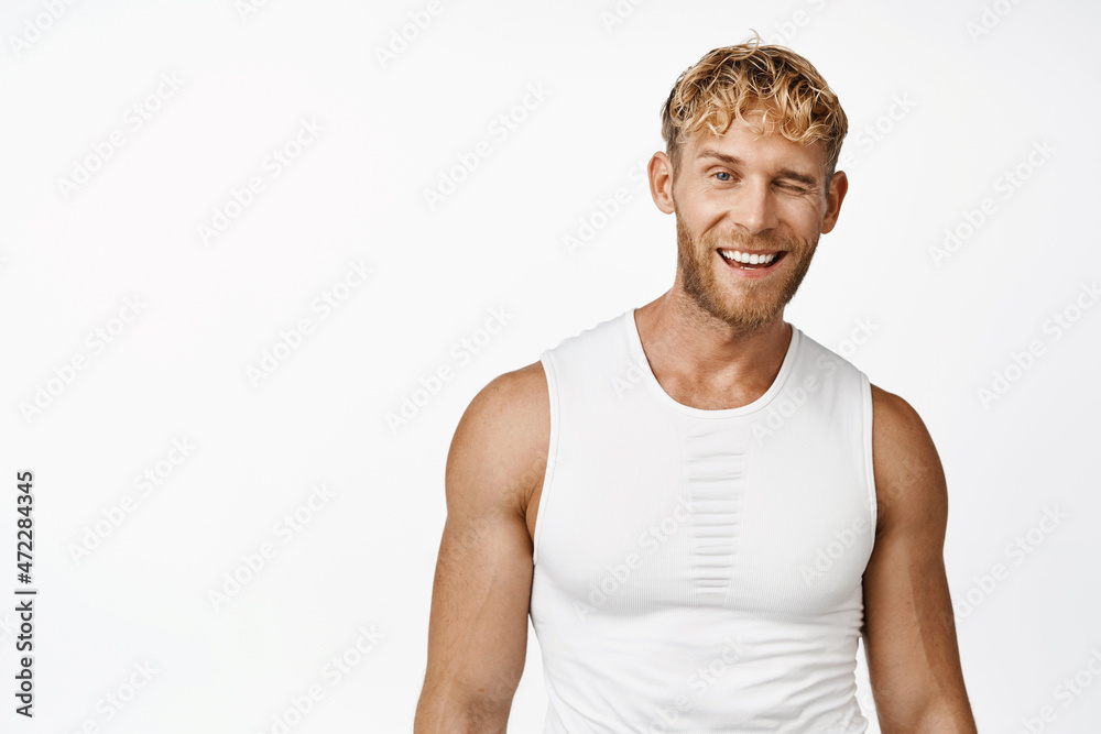 Portrait of sportsman winking and smiling, standing in workout clothes in gym, white background Portrait of sportsman winking and smiling, standing in workout clothes in gym, white background