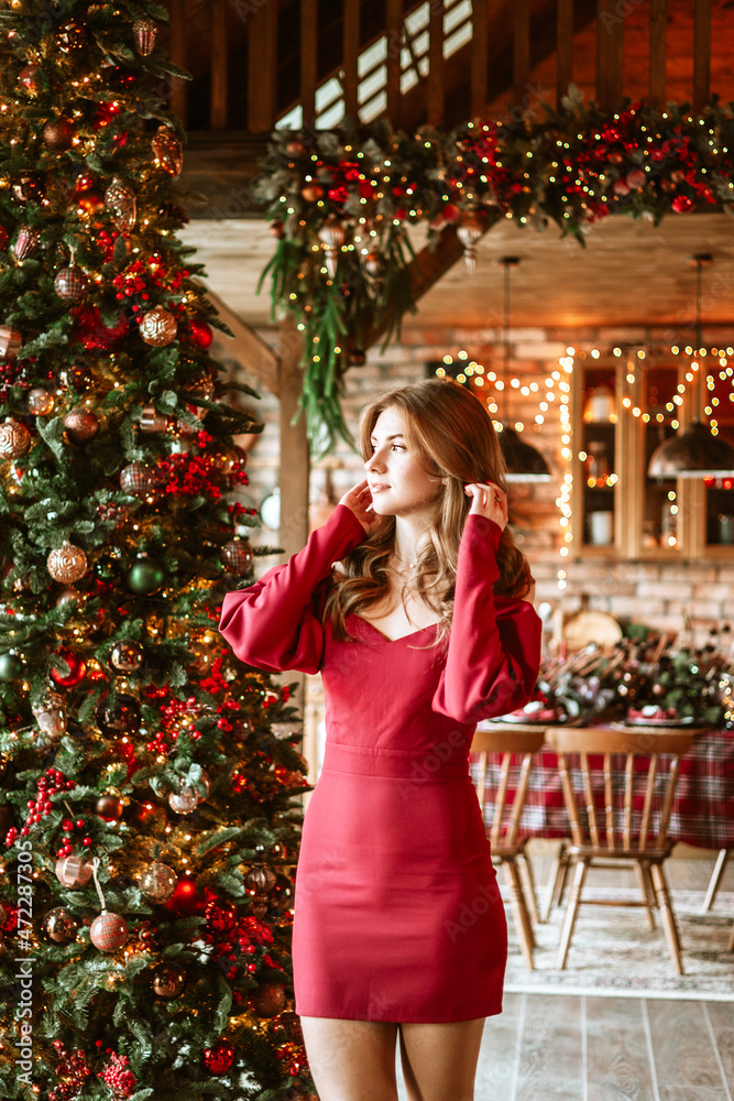 beautiful young woman in red dress stands near Christmas tree in living room decorated for celebration of Christmas and New Year in stylish interior, table set for family dinner