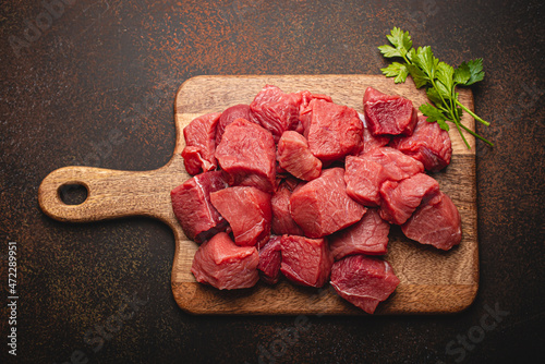 Raw beef meat chopped in cubes with bunch of fresh parsley, garlic, salt and pepper on wooden cutting board for cooking stew or meat dish on brown dark stone concrete background top view flat lay