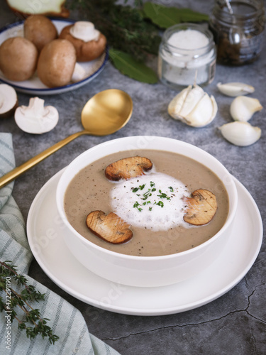A bowl of silky and creamy texture mushroom soup topped with foam mousse cream, parsley and grilled sliced mushroom. With its ingredient such thyme, garlic, onion and spoon in background 