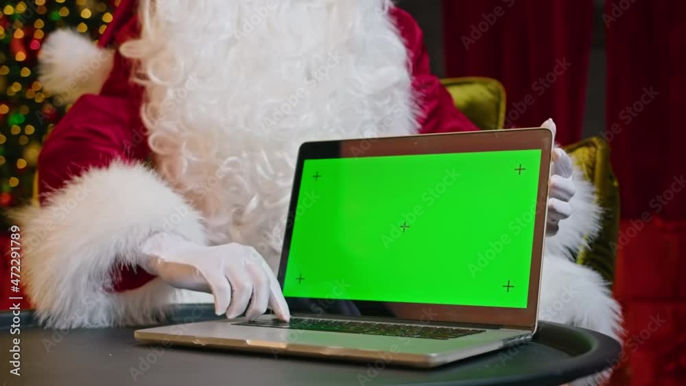 Modern Santa Claus. Cheerful Santa Claus is working on a laptop and smiling while sitting in his chair and a Christmas tree in the background. Modern Santa Claus. Cheerful Santa Claus is working on a laptop and smiling while sitting in his chair and a Christmas tree in the background.