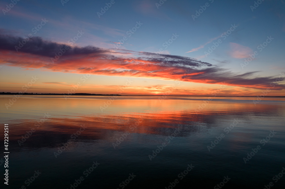 Sundown over the lake. Blue and orange sky, red sun during summer sunset. Mirror image of clouds in the water.