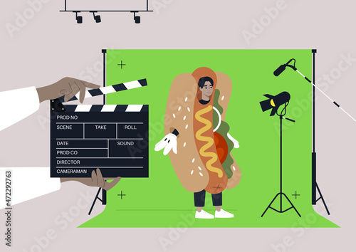 A young male Asian character wearing a hot dog costume at the commercial filming, a chroma key screen, lighting equipment, a microphone, a clapper board