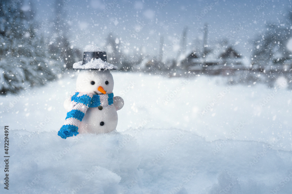 Snowman stands in a snowdrift with a village on the background in snowy evening
