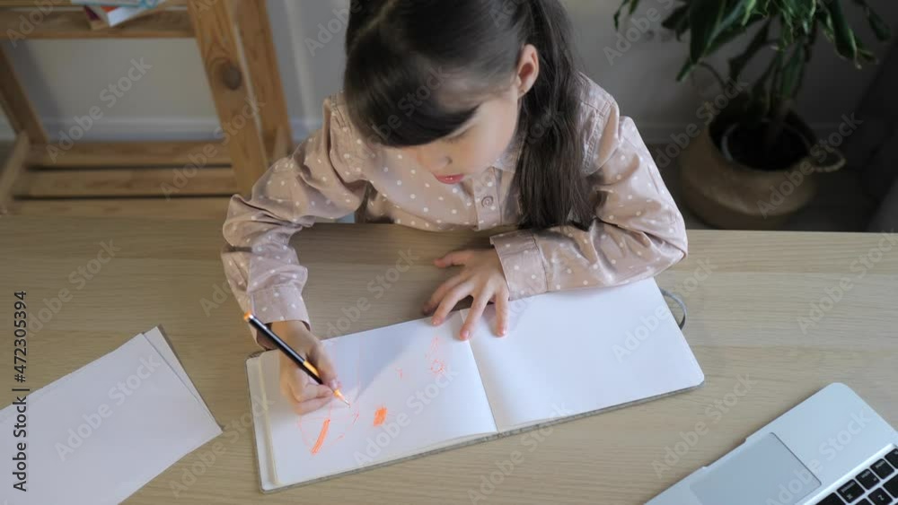 Little girl is drawing in notebook at home with laptop on desk. Online learning, homeschooling. Kid is doing homework, technology concept Little girl is drawing in notebook at home with laptop on desk. Online learning, homeschooling. Kid is doing homework, technology concept