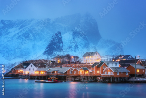 Beautiful yellow rorbuer and houses in Sarkisoy village, Lofoten islands, Norway. Winter landscape with traditional norwegian rorbuer, sea, snowy mountains in fog at night. Old fishermen's houses