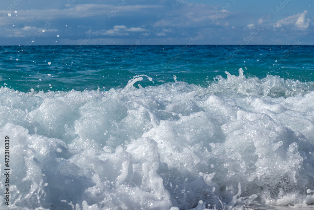 Obraz premium Shining waves splash with white foam drops. Blue vivid sea and epic clouds on coast of Lefkada island in Greece. Summer travel to Ionian Sea