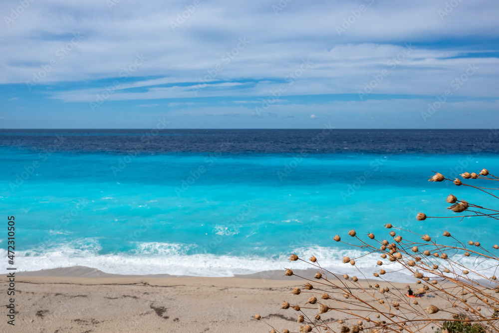 Obraz premium Azure vibrant waves with dry wild flowers and blue sky on sunny coast of Greek island. Sandy beach in Greece. Summer nature vacation travel to Ionian Sea