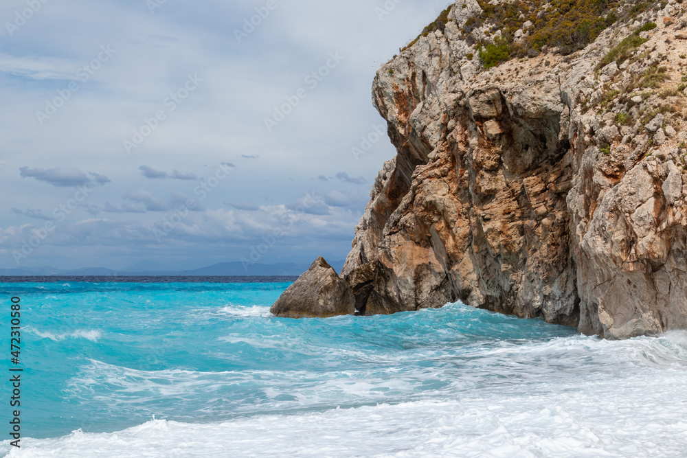 Azure stormy waves with white foam breaking rocky coast. Mylos beach with dramatic cloudy sky on Lefkada island in Greece. Summer nature travel to Ionian Sea Azure stormy waves with white foam breaking rocky coast. Mylos beach with dramatic cloudy sky on Lefkada island in Greece. Summer nature travel to Ionian Sea