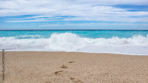 Fototapeta Naklejka Na Ścianę i Meble -  Azure vibrant waves hitting shore with blue sky on sunny coast of Greek island. Sandy beach with footprints in Greece. Summer travel to Ionian Sea