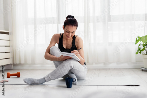 Young woman writing in her notebook. Sitting at home by the window on a yoga mat after exercise. Personal growth resolutions 