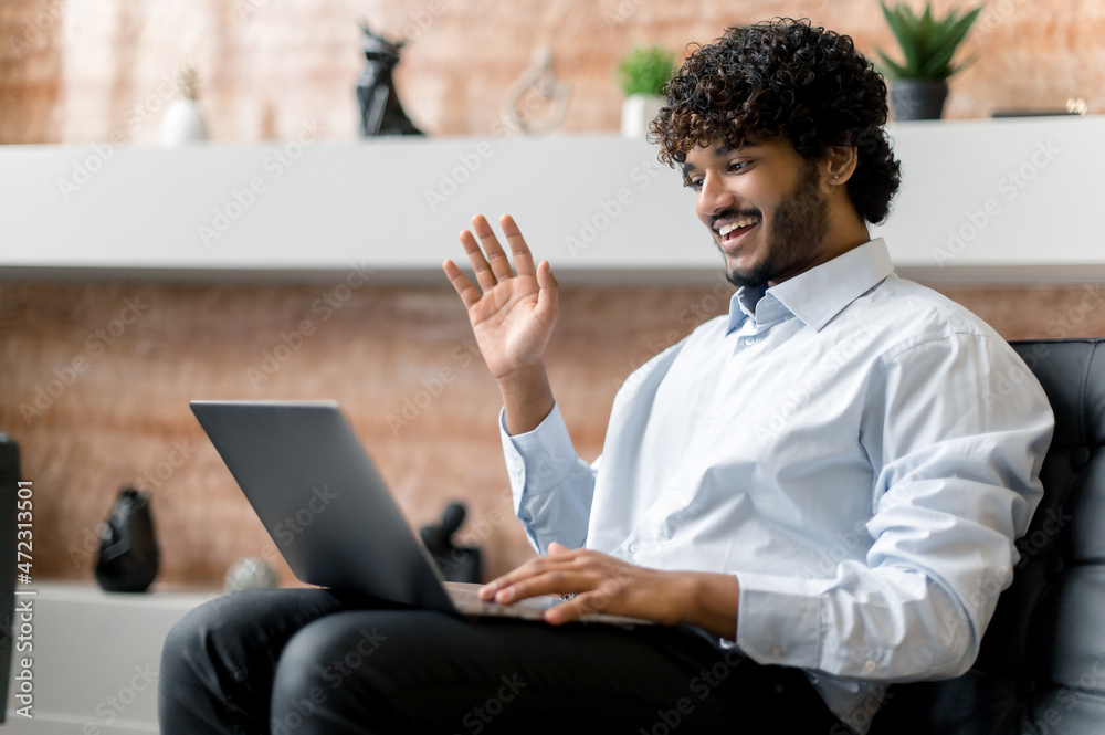 Online video communication. Positive Indian guy, sit on a armchair, uses a laptop, talks on a video conference with friends, colleagues or family, chatting on social networks, greets the interlocutor Online video communication. Positive Indian guy, sit on a armchair, uses a laptop, talks on a video conference with friends, colleagues or family, chatting on social networks, greets the interlocutor