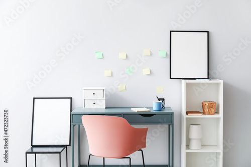 Modern workplace with shelving unit and blank frames near light wall