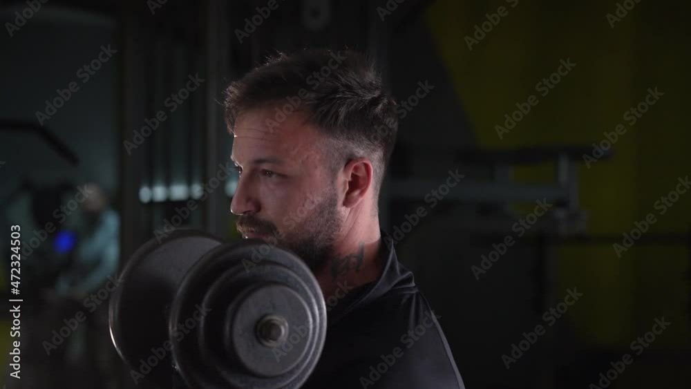 Side view portrait of young caucasian man male athlete bodybuilder training at the gym workout using dumbbells biceps curls wearing black shirt dark hair and beard standing weight lifting Side view portrait of young caucasian man male athlete bodybuilder training at the gym workout using dumbbells biceps curls wearing black shirt dark hair and beard standing weight lifting