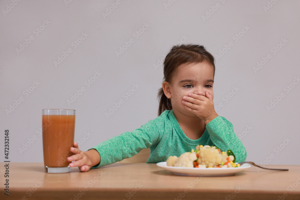 Cute little girl covering mouth and refusing to drink juice at table on grey background Cute little girl covering mouth and refusing to drink juice at table on grey background