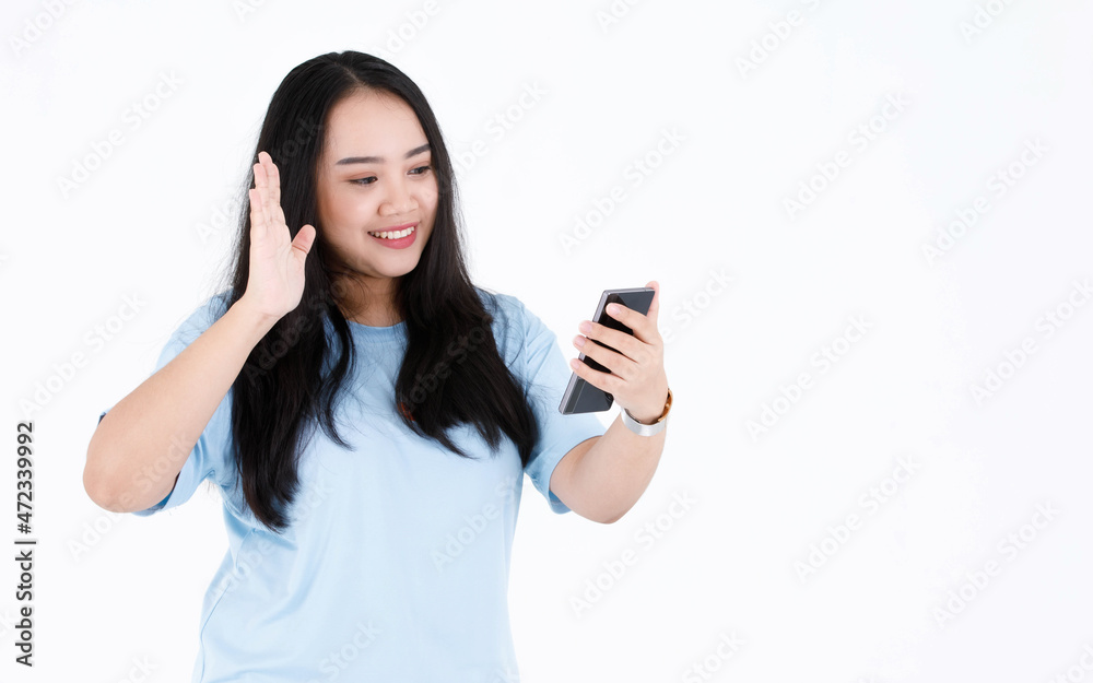 Studio shot of Asian chubby plump long black hair female model in blue casual t shirt and wristwatch smiling holding smartphone waving hand greeting say hi hello on video call on white background Studio shot of Asian chubby plump long black hair female model in blue casual t shirt and wristwatch smiling holding smartphone waving hand greeting say hi hello on video call on white background