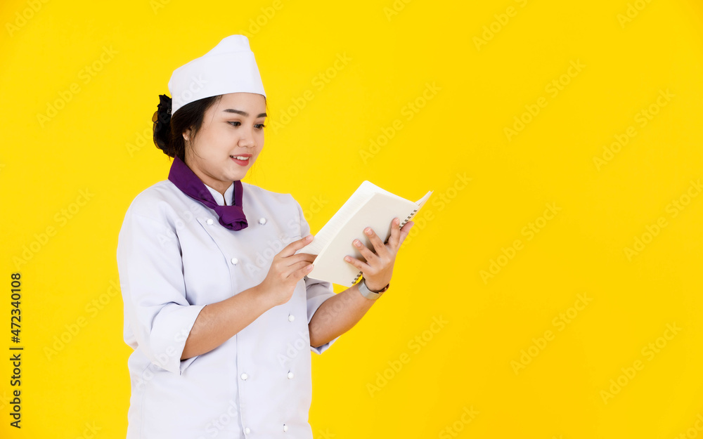 Portrait studio shot of Asian professional female executive chef in white cooking uniform with hat and scarf standing smiling holding learning new recipes from notebook in hands on yellow background Portrait studio shot of Asian professional female executive chef in white cooking uniform with hat and scarf standing smiling holding learning new recipes from notebook in hands on yellow background