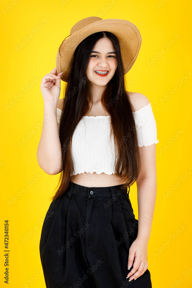 Portrait studio shot of Asian young beautiful sexy long black hair female model wearing crop top outfit dental care braces standing smiling holding hand touching adjusting hat on yellow background Portrait studio shot of Asian young beautiful sexy long black hair female model wearing crop top outfit dental care braces standing smiling holding hand touching adjusting hat on yellow background