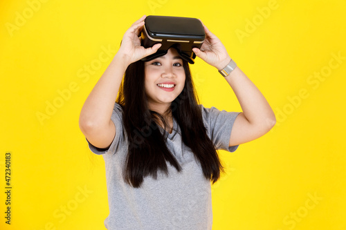 Portrait studio shot of Asian happy young chubby plump long black hair female gamer model in casual outfit smiling wearing big virtual reality vr camera goggles console game on yellow background