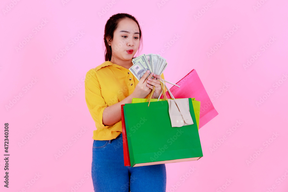 Fototapeta premium Portrait studio shot of Asian young rich wealthy female shopaholic shopper in casual outfit smiling holding hundred dollars fan shopping bags on pink background