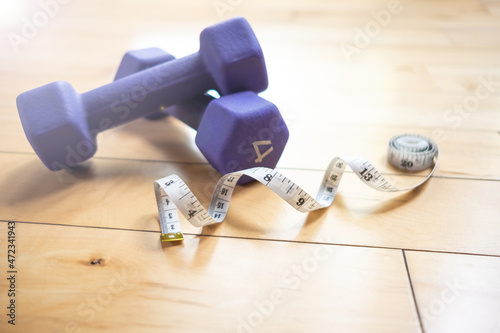 A white tape measure laying on a hardwood floor next to two small purple dumbbells inside a gym