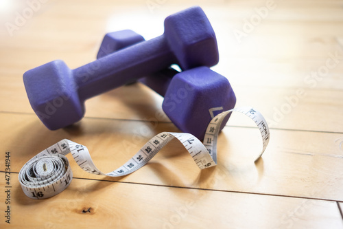 A white tape measure laying on a hardwood floor next to two small purple dumbbells inside a gym