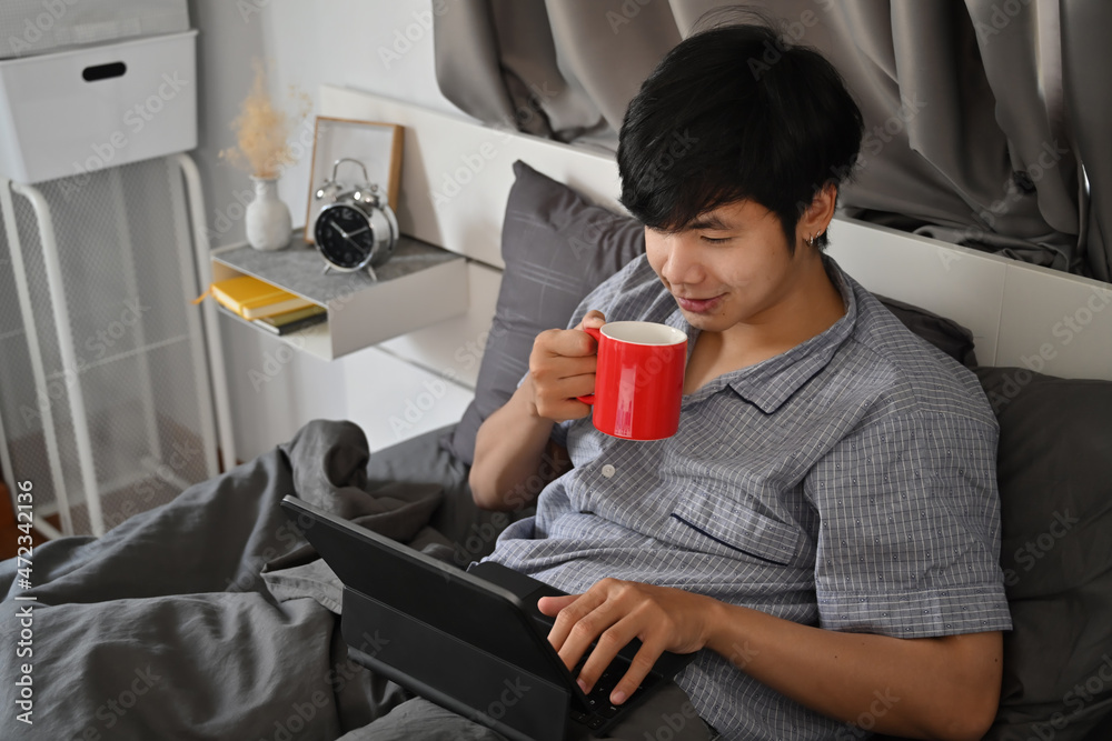 Happy asian man drinking coffee and surfing internet with computer tablet on bed.