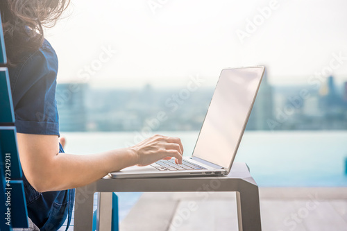 Cropped image of Woman typing on laptop while sitting near pool and Blurred background skyscraper with Beautiful sky and bright sunlight. Copy space for text
