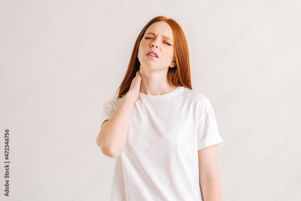 Portrait of suffering upset young woman with grimace of pain, touching neck feeling pain and numbness, worried about muscle tension, osteochondrosis, standing on white isolated background in studio. Portrait of suffering upset young woman with grimace of pain, touching neck feeling pain and numbness, worried about muscle tension, osteochondrosis, standing on white isolated background in studio.