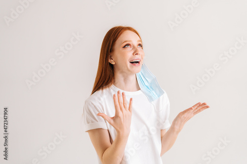 Studio portrait of happy cheerful young woman in casual t-shirt taking off disposable protective face mask, standing on white isolated background. Flu epidemic, dust allergy, protection against virus