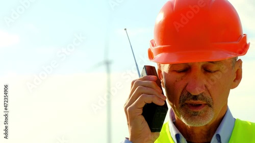 Windmill rotates producing energy under cloudy sky. Engineer in orange helmet gives information via radio set at construction site closeup photo