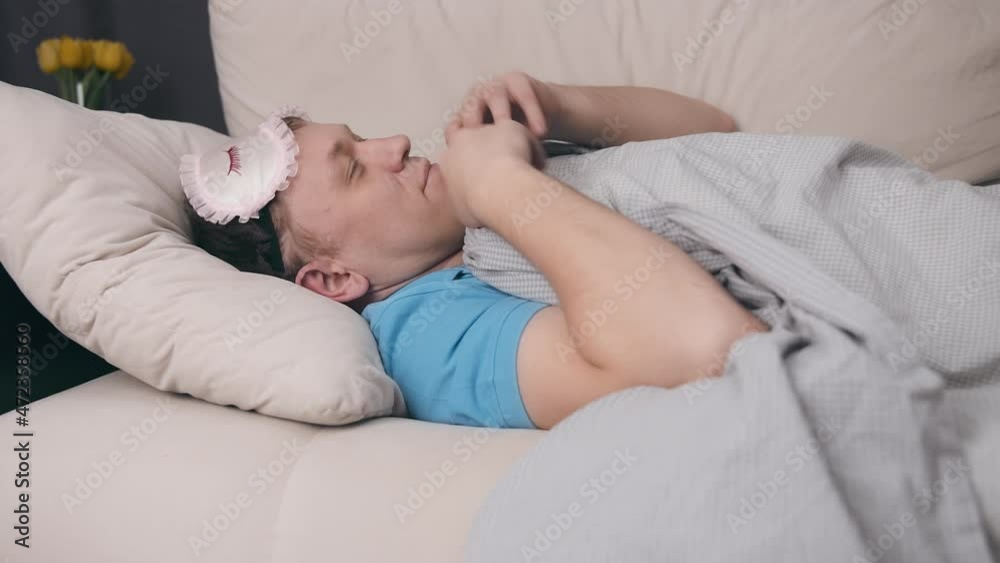 Man in a mask lies on the sofa covered with a blanket, puts a mask on his eyes and falls asleep Man in a mask lies on the sofa covered with a blanket, puts a mask on his eyes and falls asleep