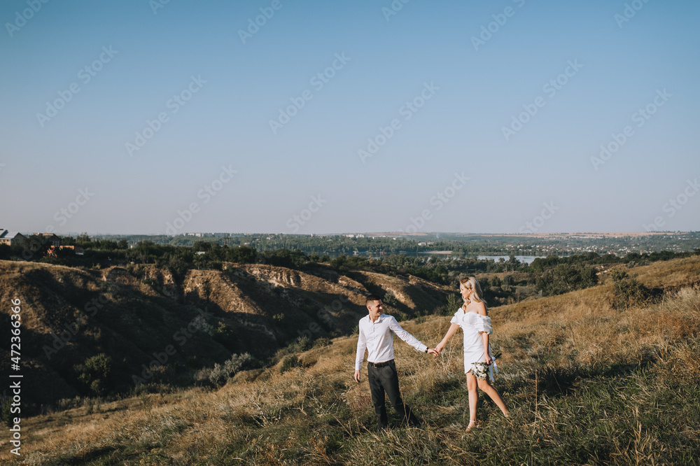A young groom and a beautiful blonde bride in a white dress are walking, holding hands, in nature on the background of hills and cliffs in autumn. Wedding photography.
