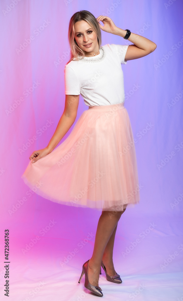 Naklejka premium beautiful woman portrait in studio posing. A girl in a white blouse, a T-shirt and a pink, peach pleated skirt laughs, spins, dances. colored, crimson purple background. holiday, new year, birthday