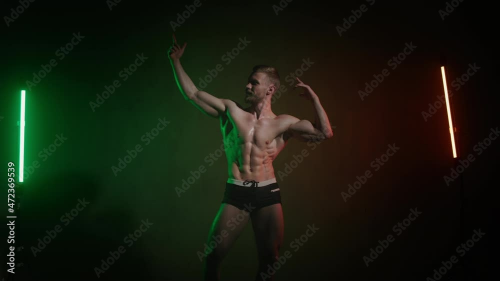 A sportsman with a bare body is posing and raises his hands for showing muscles. An orange and green light is shining on him. 4K A sportsman with a bare body is posing and raises his hands for showing muscles. An orange and green light is shining on him. 4K