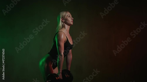The fitness model is standing and doing a dumbbells deadlift. She is bending her arm with a dumbbell. She is bending down. An orange and green light is shining on her. 4K 50fps
