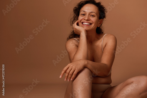 Self-confident beautiful Hispanic woman in beige underwear, smiles toothy smile looking at camera. Body positivity, self-acceptance and body love concept photo