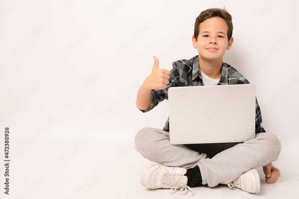 Smiling cute boy wearing plaid shirt sitting on floor holding laptop computer with thumb up isolated over white background. Smiling cute boy wearing plaid shirt sitting on floor holding laptop computer with thumb up isolated over white background.