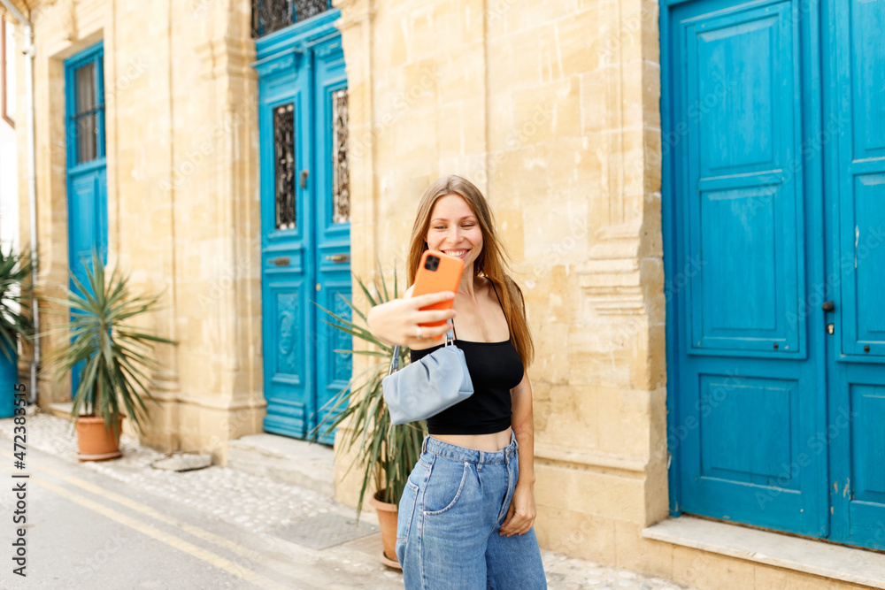 Young beautiful girl in denim jeans and purse handbag makes selfie against the background of the Cyprus old town.