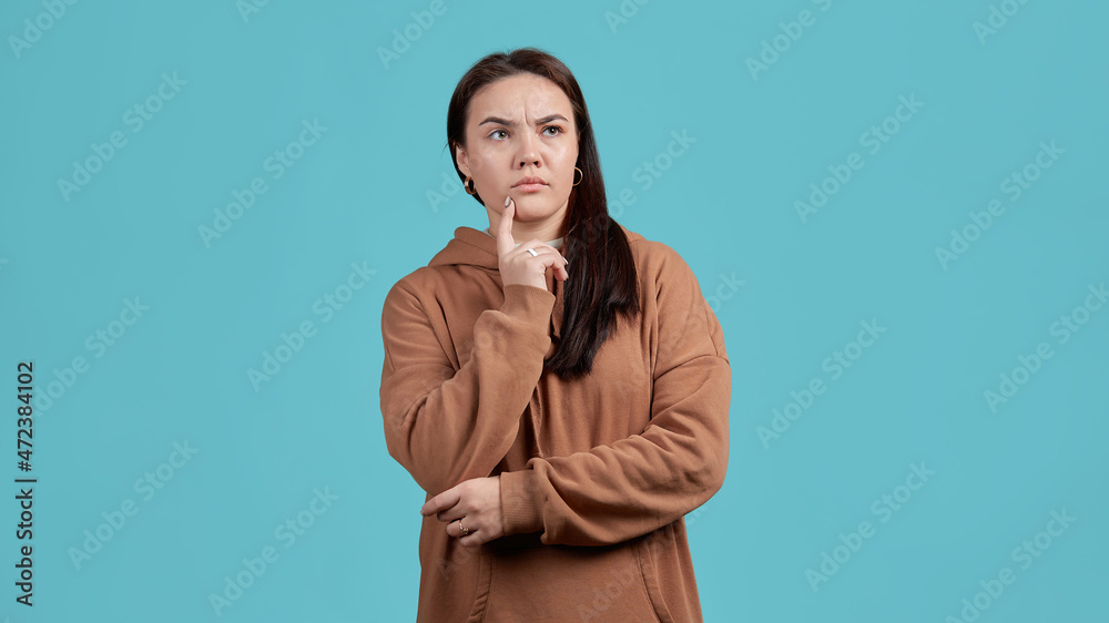 A young woman in a brown hoodie, standing dreamily on a blue isolated background. The concept of thoughtfulness, comprehension, problem solving in the head. A young woman in a brown hoodie, standing dreamily on a blue isolated background. The concept of thoughtfulness, comprehension, problem solving in the head.