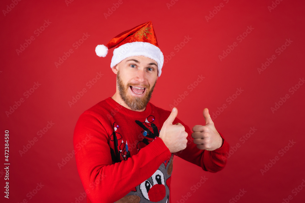 Red-bearded man in a santa hat and a christmas sweater on a red background cheerfully smiles thumbs up Red-bearded man in a santa hat and a christmas sweater on a red background cheerfully smiles thumbs up