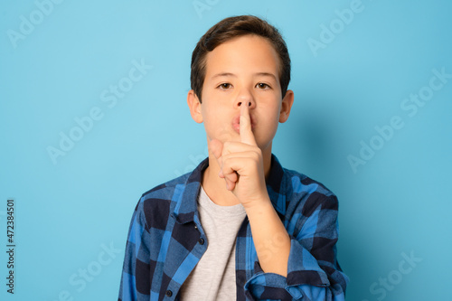 Kid boy asking to be quiet with finger on lips, isolated on blue background.