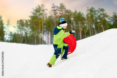 childhood, sledging and season concept - happy little boy with snow saucer sled outdoors in winter over snowy forest or park background