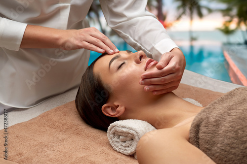 wellness, beauty and relaxation concept - beautiful young woman lying with closed eyes and having face and head massage at spa over tropical beach background in french polynesia