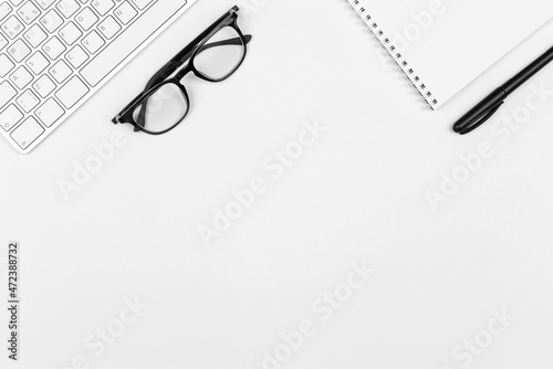 Keyboard, black eyeglasses, notepad, pen on white background. Minimal business concept. Office supplies on table. Monochrome workspace. Copy space, flat lay, top view.