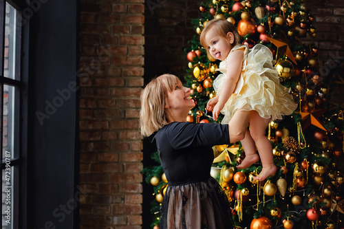 A blonde mother in a black dress and a little daughter near a Christmas tree with golden balls. A woman holds a girl in her arms.