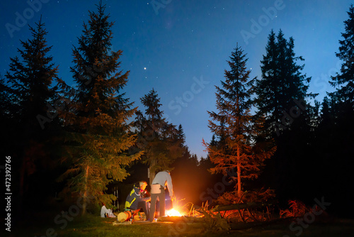 Bonfire in night forest and people near fire under night dark sky with stars
