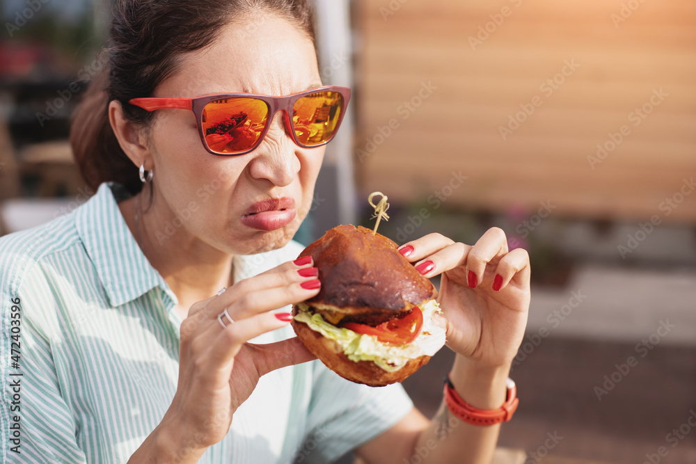 Worried and anxious woman in a street cafe tries a disgusting or spoiled burger. The concept of harmful fast food and excessive calories Worried and anxious woman in a street cafe tries a disgusting or spoiled burger. The concept of harmful fast food and excessive calories