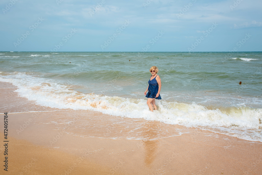 Happy mature woman enjoying on the sea beach, walking on the waves. An elderly woman in a dark blue swimsuit on the seashore. Happy mature woman enjoying on the sea beach, walking on the waves. An elderly woman in a dark blue swimsuit on the seashore.